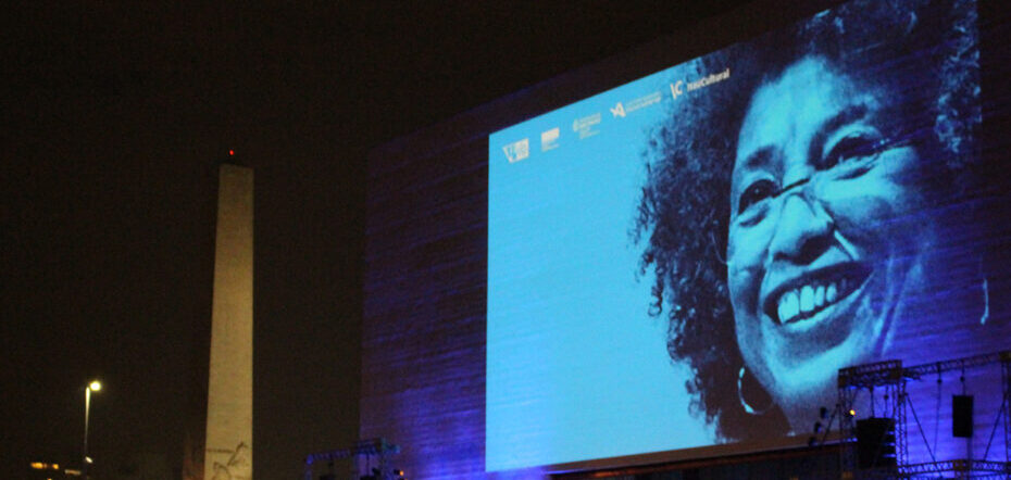 Foto de um palco com uma foto de Angela Davis no telão. Ela é negra, usa óculos de grau e sorri