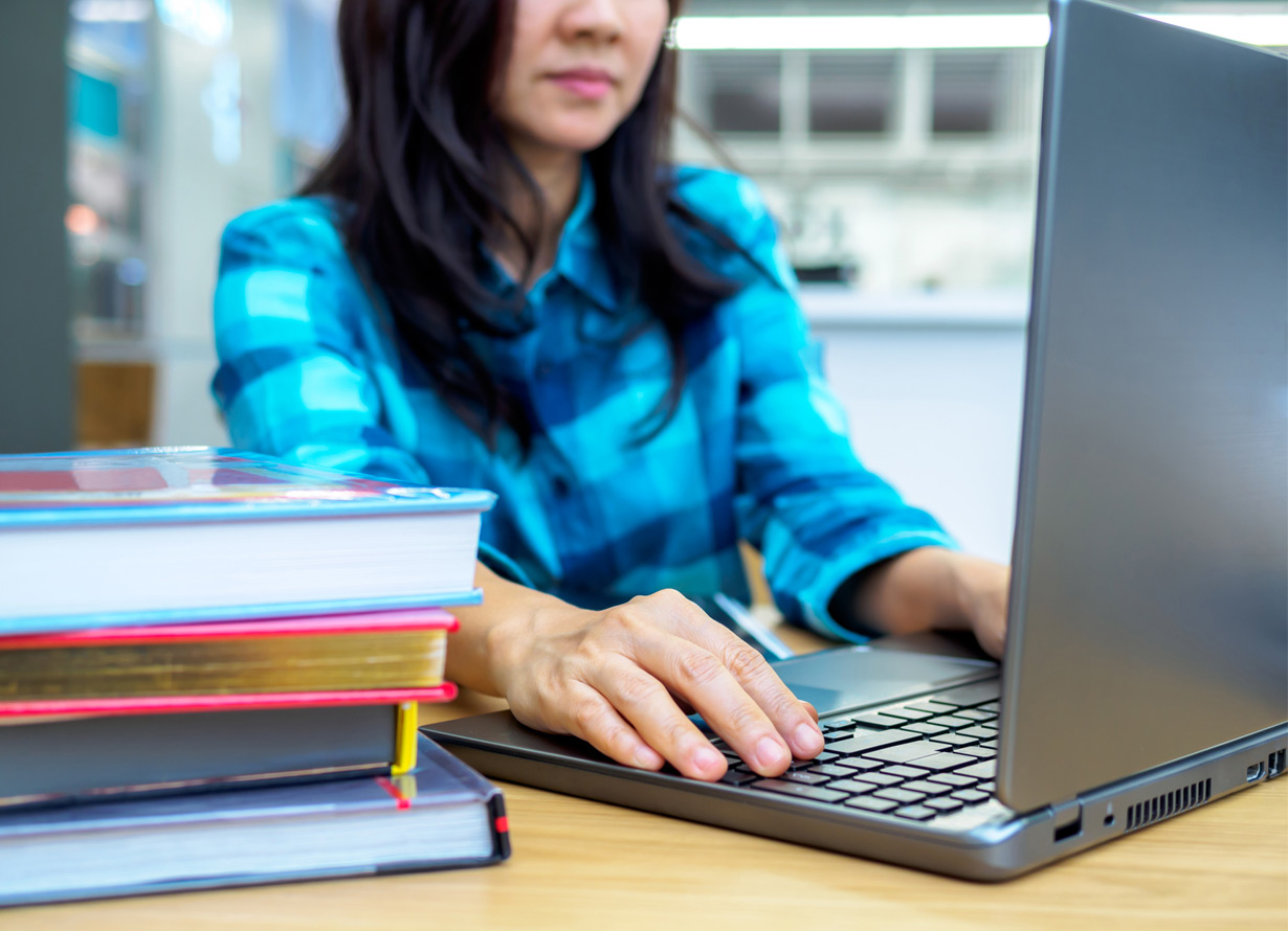 Foto de uma mulher mexendo em um notebook que está sobre uma mesa. Ao lado, há diversos livros empilhados