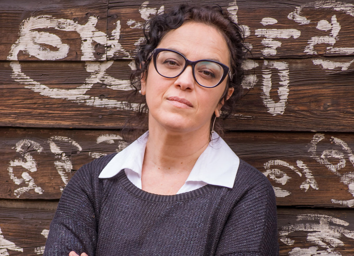 Foto de uma mulher de cabelos escuros cacheados e óculos de grau. Ela está de braços cruzados e apoiada em uma madeira com desenhos feitos com tinta branca