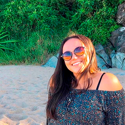 Foto de uma mulher de cabelos castanhos e lisos sorrindo na praia. Atrás dela, há pedras e árvores.