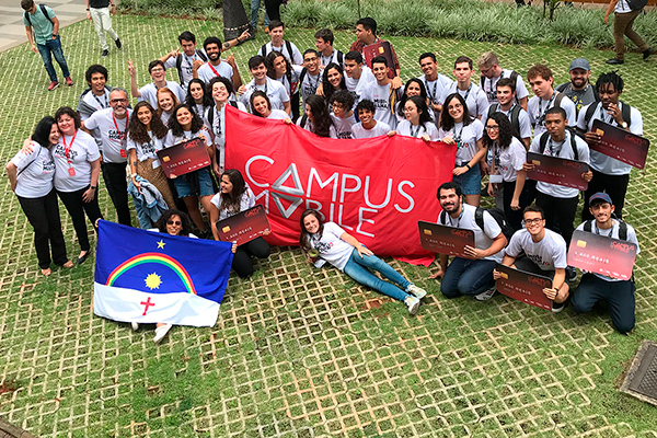 Foto tirada de cima de um grupo diverso de pessoasem um local gramado. Eles sorriem e alguns seguram uma faixa vermelha do Campus Mobile e réplicas de cartões de crédito em tamanho grande nas mãos
