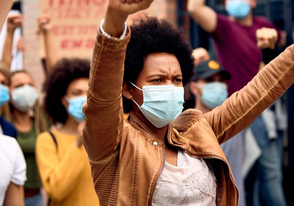 Foto de uma mulher negra usando máscara de proteção no rosto com os braços levantados em sinal de protesto. Ao fundo, há pessoas segurando cartazes e com os braços levantados.