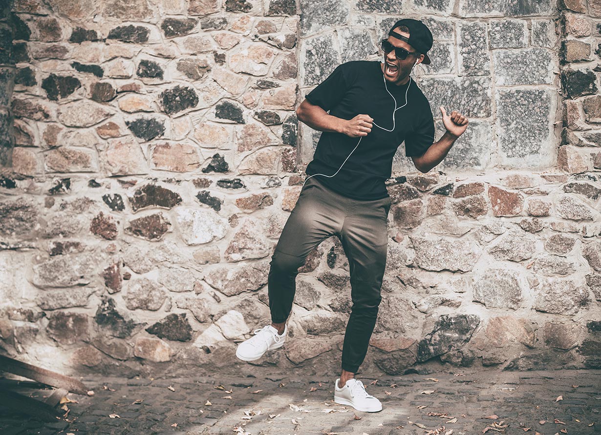 Foto de um homem negro em pé usando fones de ouvido. Ele está com um pé levantado e com a boca aberta como se estivesse dançando (crédito: g-stockstudio – iStock)