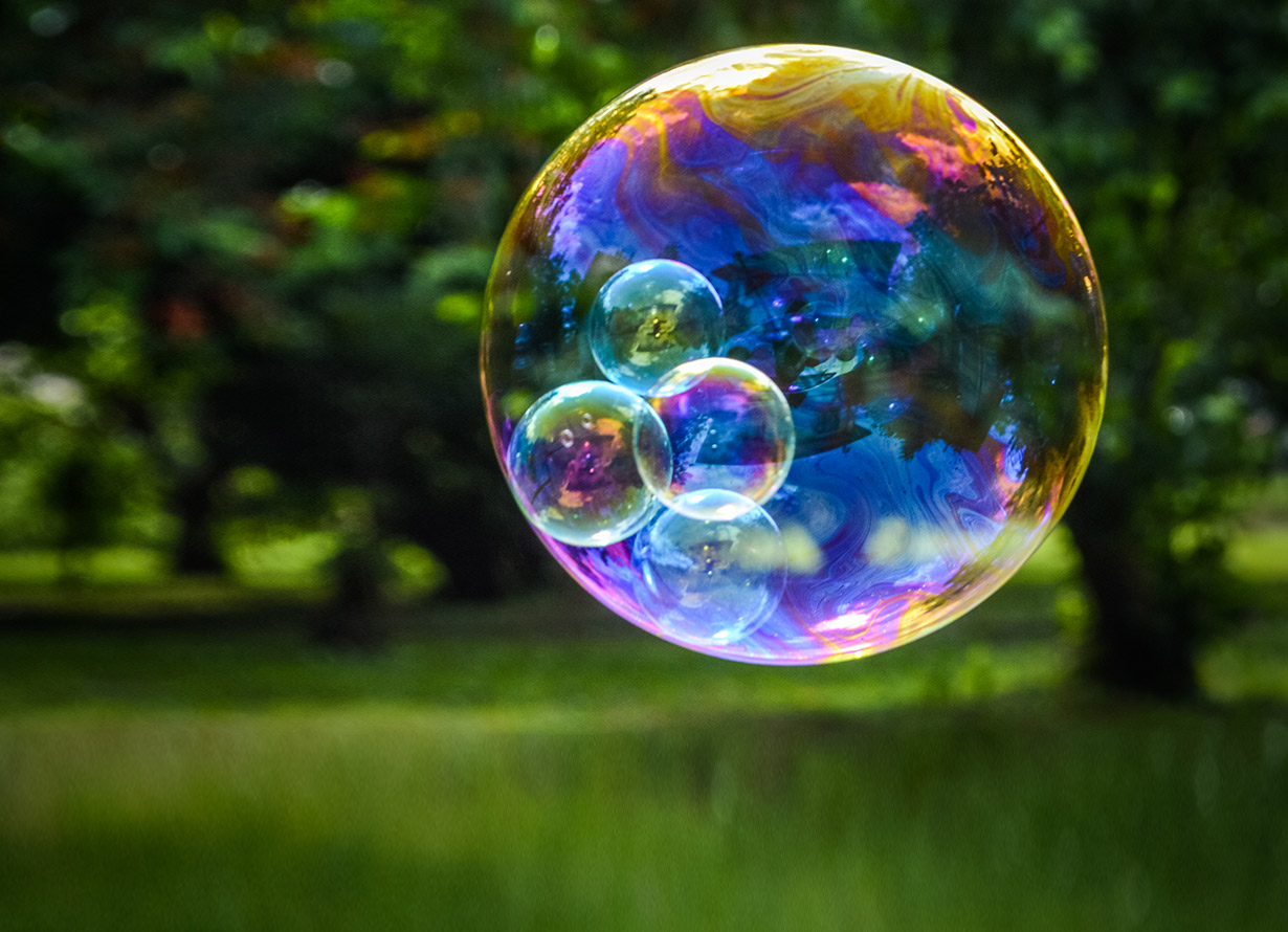 Foto de uma grande bolha de sabão com bolhas menores dentro dela. Ao fundo, há uma área verde arborizada